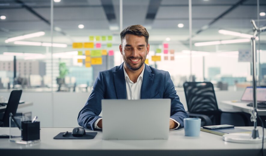 Ein Mann arbeitet lächelnd an seinem Laptop in einem modernen Büro.