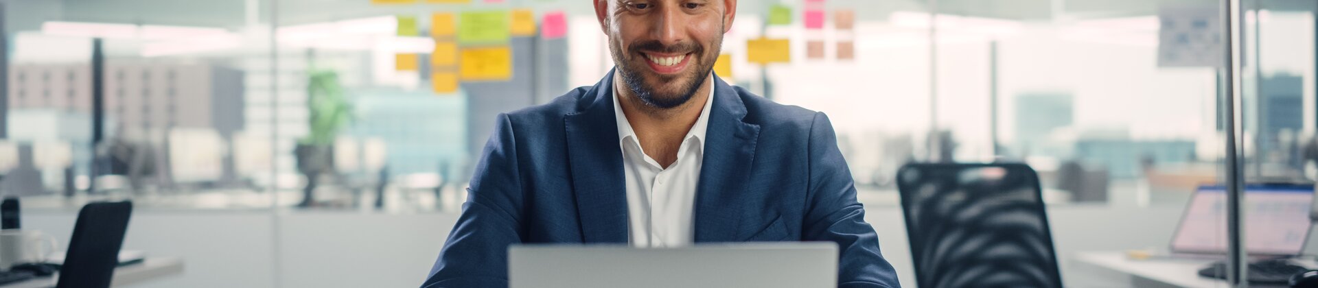 Ein Mann arbeitet lächelnd an seinem Laptop in einem modernen Büro.