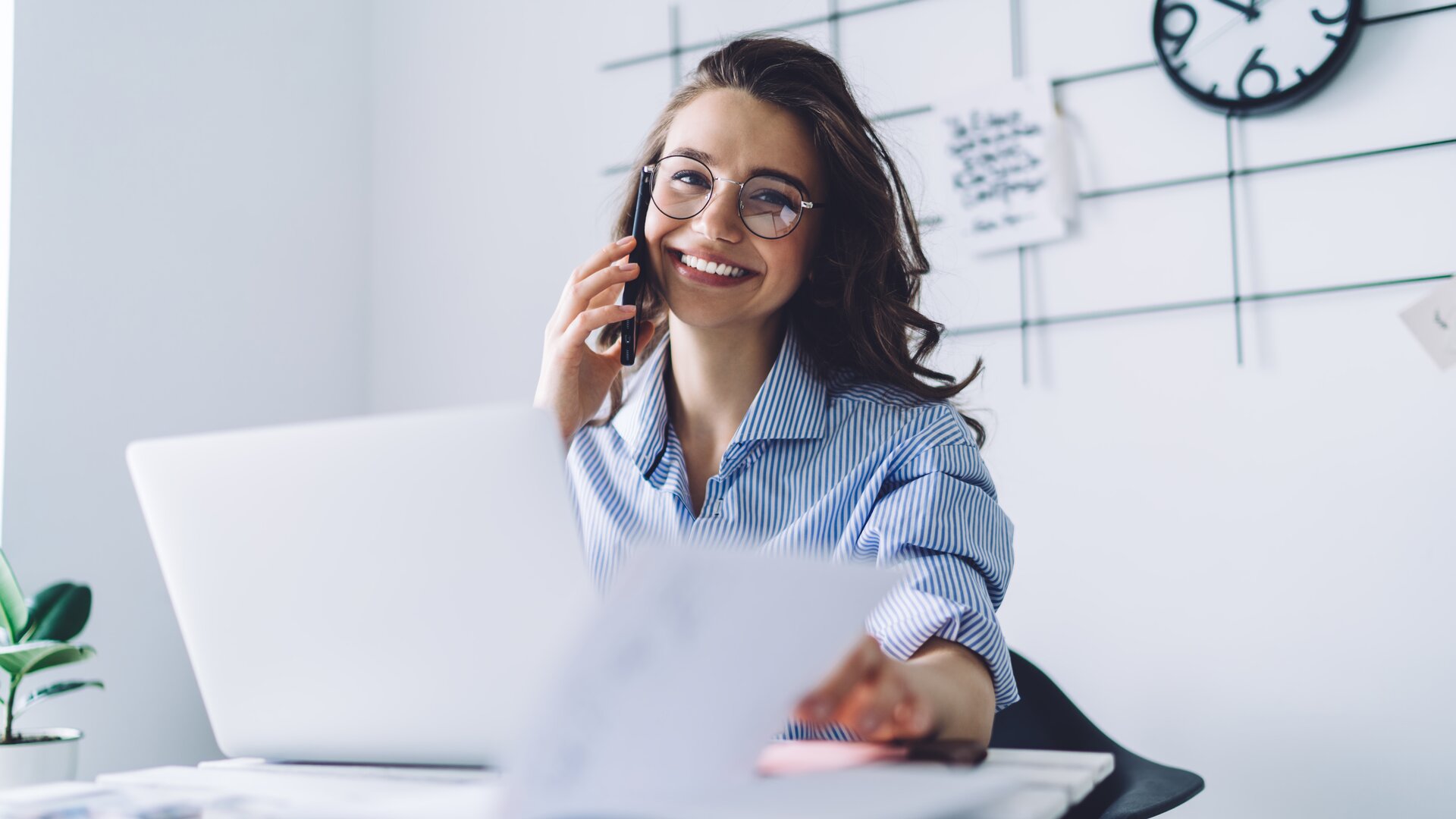 Eine lächelnde Frau telefoniert am Schreibtisch und arbeitet gleichzeitig an Unterlagen vor ihrem Laptop.
