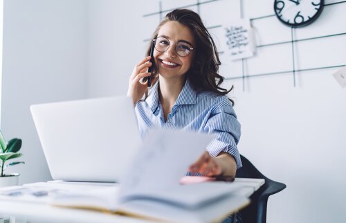 Eine lächelnde Frau telefoniert am Schreibtisch und arbeitet gleichzeitig an Unterlagen vor ihrem Laptop.