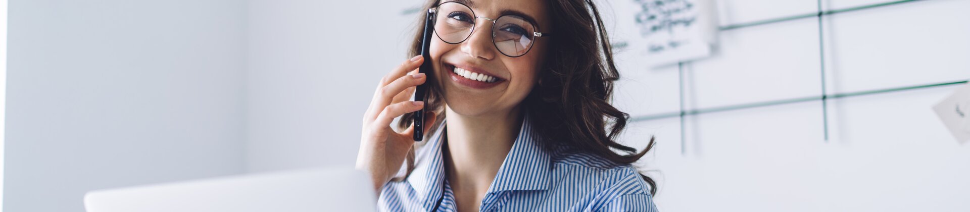 Eine lächelnde Frau telefoniert am Schreibtisch und arbeitet gleichzeitig an Unterlagen vor ihrem Laptop.