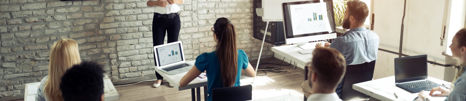 Eine Frau präsentiert Geschäftsgrafiken vor einer aufmerksam zuhörenden Gruppe in einem Seminarraum.
