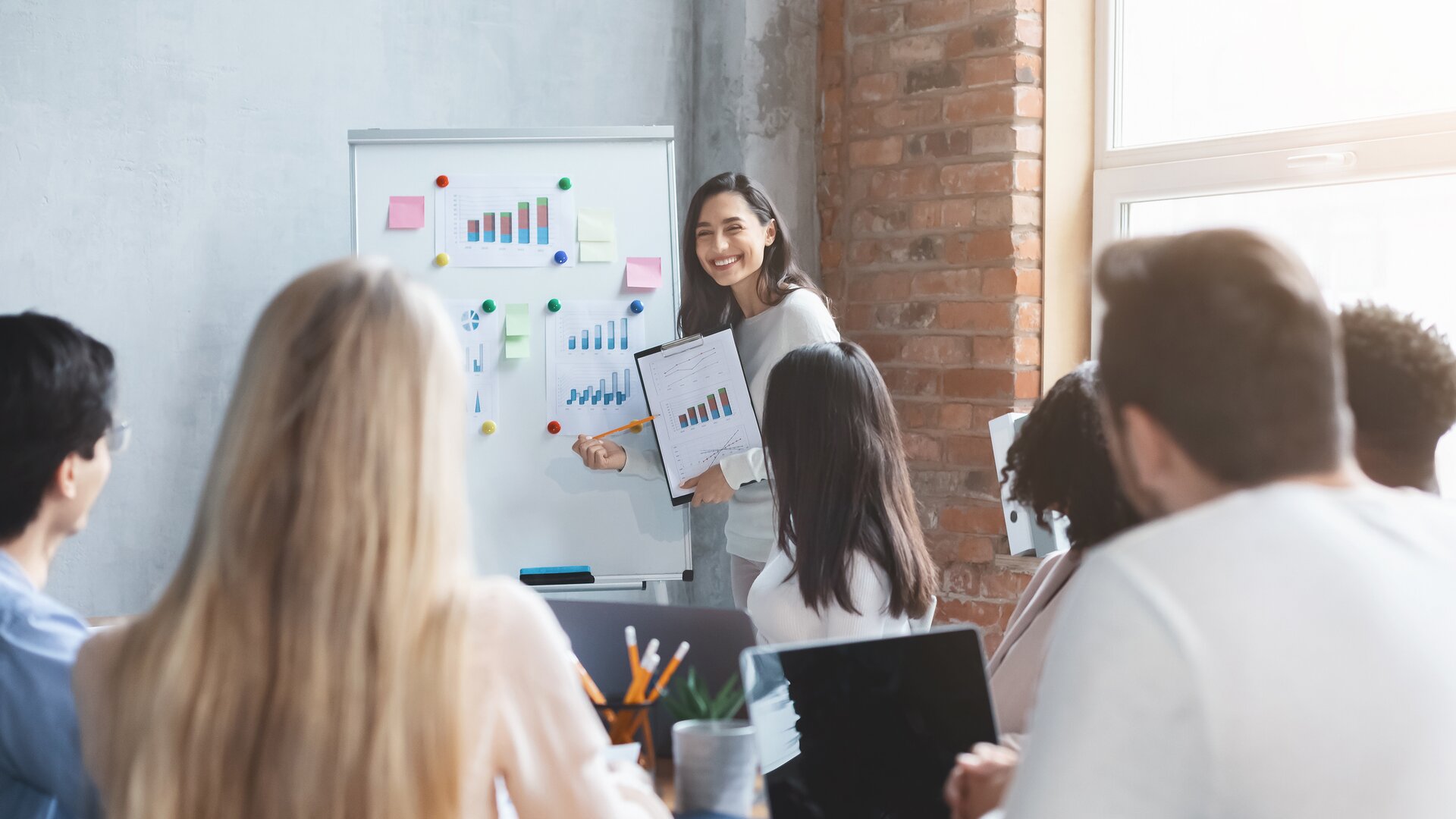 Eine Frau präsentiert ihrem Team gut sichtbar Diagramme und Ergebnisse an einem Whiteboard.