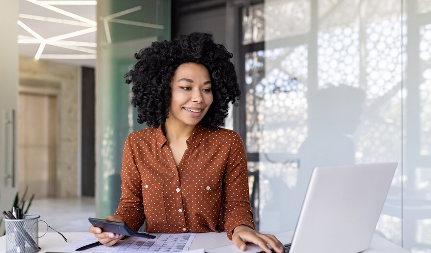Das Bild zeigt eine Person, die konzentriert am Schreibtisch arbeitet und dabei sowohl einen Laptop als auch einen Taschenrechner benutzt.