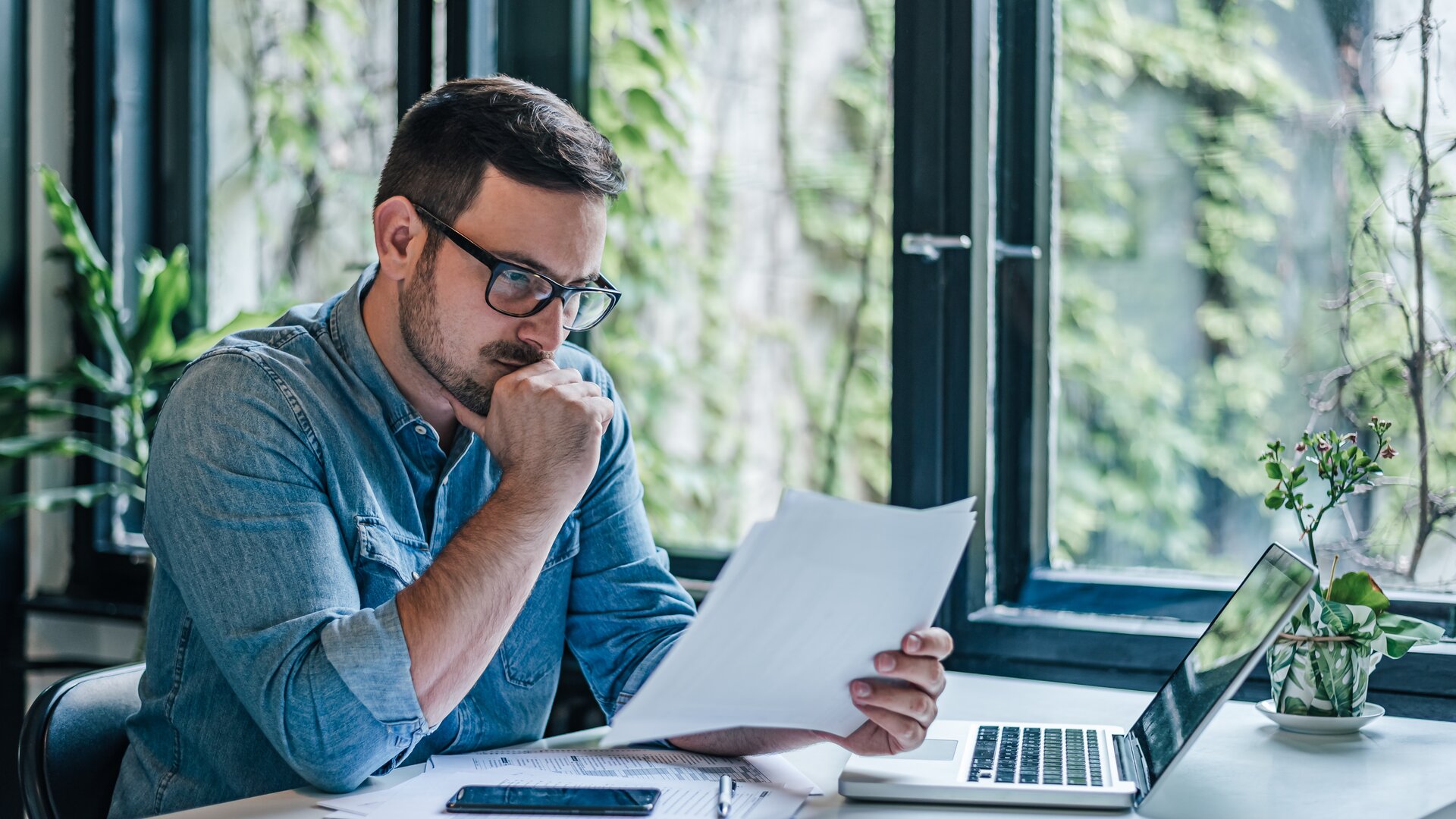 Ein Mann arbeitet konzentriert an seinem Schreibtisch und prüft aufmerksam Dokumente vor einem geöffneten Laptop.