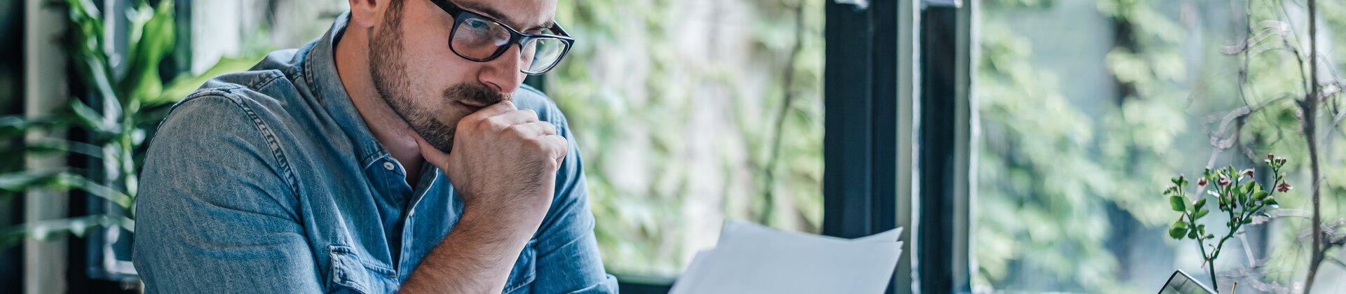 Ein Mann arbeitet konzentriert an seinem Schreibtisch und prüft aufmerksam Dokumente vor einem geöffneten Laptop.