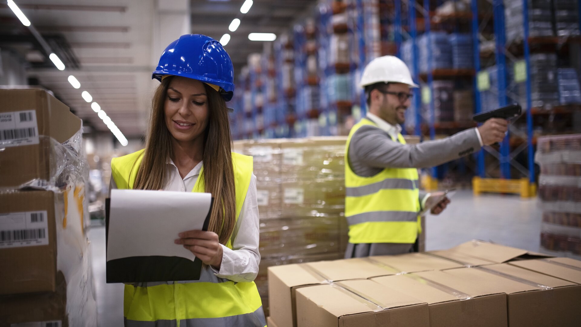 Eine Mitarbeiterin prüft Unterlagen, während ein Kollege im Lager Waren mit einem Scanner erfasst.