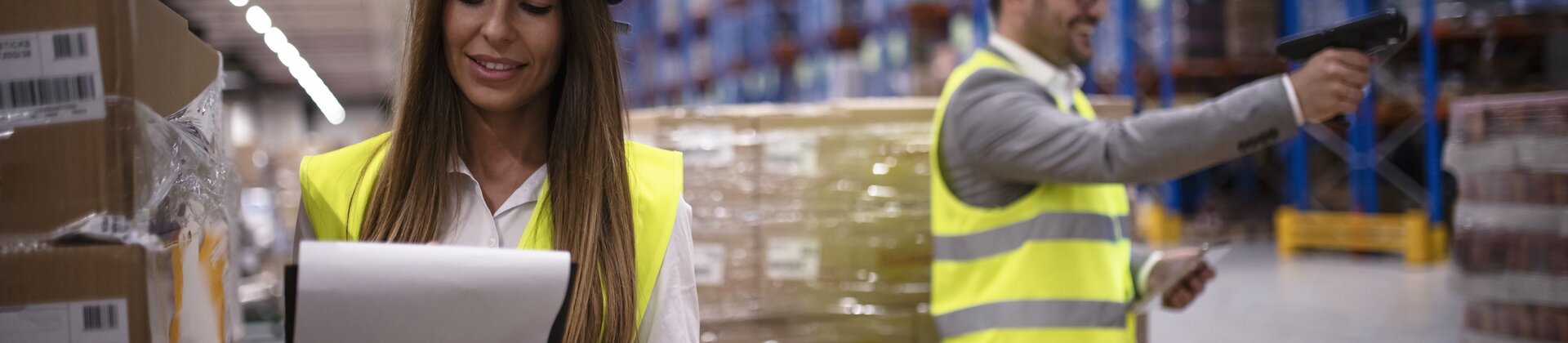 Eine Mitarbeiterin prüft Unterlagen, während ein Kollege im Lager Waren mit einem Scanner erfasst.