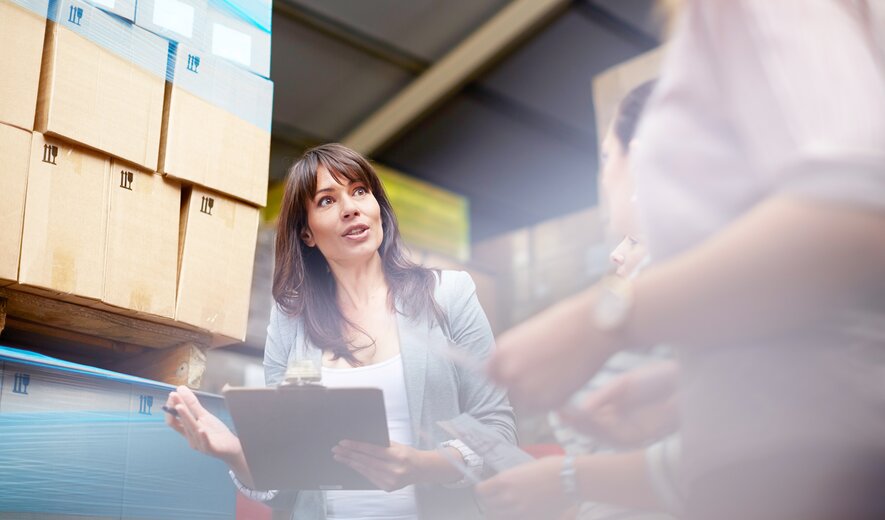 Eine Mitarbeiterin überprüft mit einem Klemmbrett Warenbestände in einem Lager und spricht dabei mit Kolleginnen.