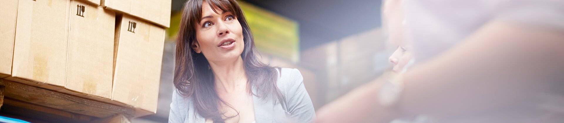 Eine Mitarbeiterin überprüft mit einem Klemmbrett Warenbestände in einem Lager und spricht dabei mit Kolleginnen.