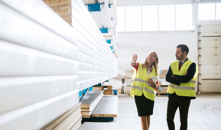 Eine Mitarbeiterin zeigt einem Kollegen in einer Lagerhalle verschiedene Materialien oder Bestände.