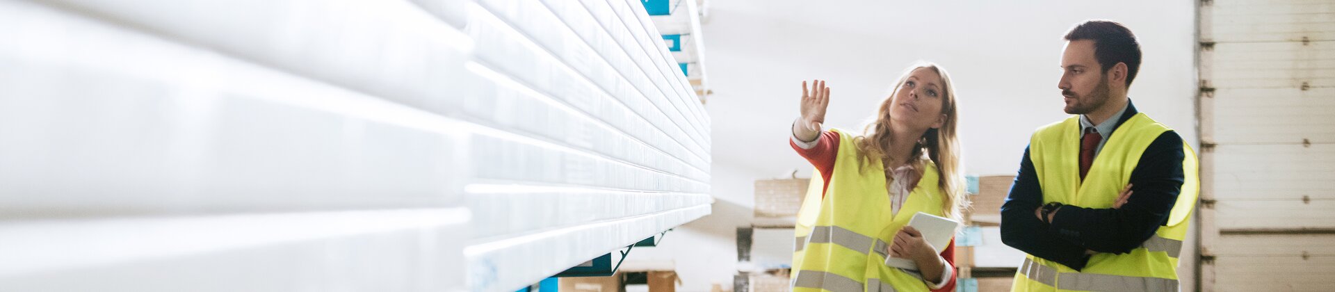 Eine Mitarbeiterin zeigt einem Kollegen in einer Lagerhalle verschiedene Materialien oder Bestände.