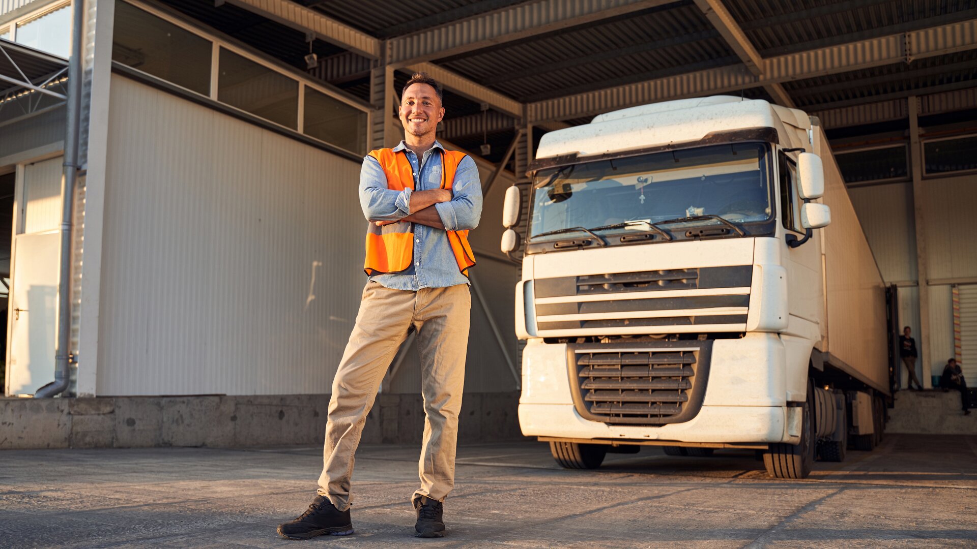 Ein Mann in Warnweste steht selbstbewusst vor einem großen Lkw auf einem Betriebshof.
