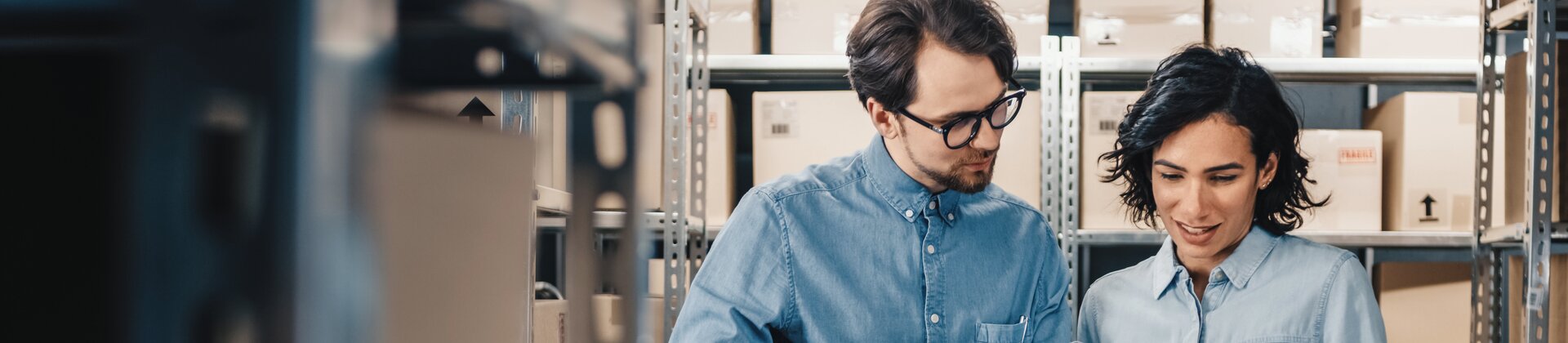 Zwei Mitarbeitende in einem Lager prüfen gemeinsam per Tablet den Bestand, während einer von ihnen einen großen Karton trägt.