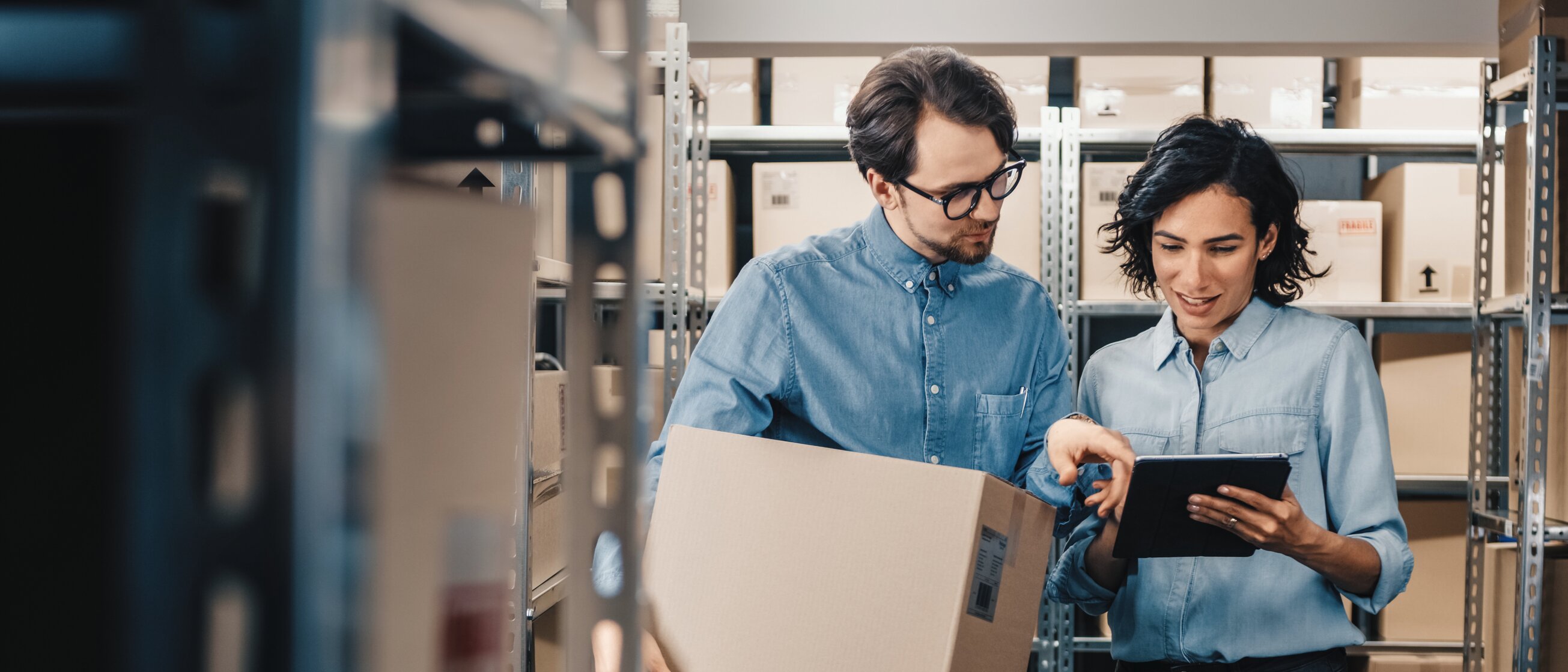 Zwei Mitarbeitende in einem Lager prüfen gemeinsam per Tablet den Bestand, während einer von ihnen einen großen Karton trägt.