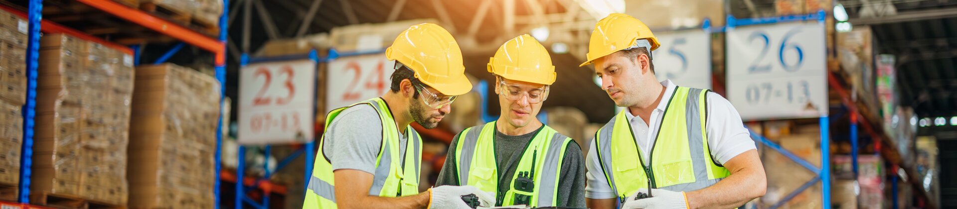 Drei Lagerarbeiter in Schutzwesten und Helmen besprechen gemeinsam Informationen auf einem Tablet mitten in einem großen Warenlager.