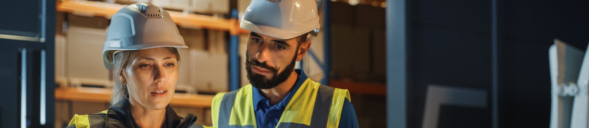 Zwei Lagerarbeiter/-innen in Warnwesten und Helmen prüfen gemeinsam Informationen auf einem Tablet in einer Halle.
