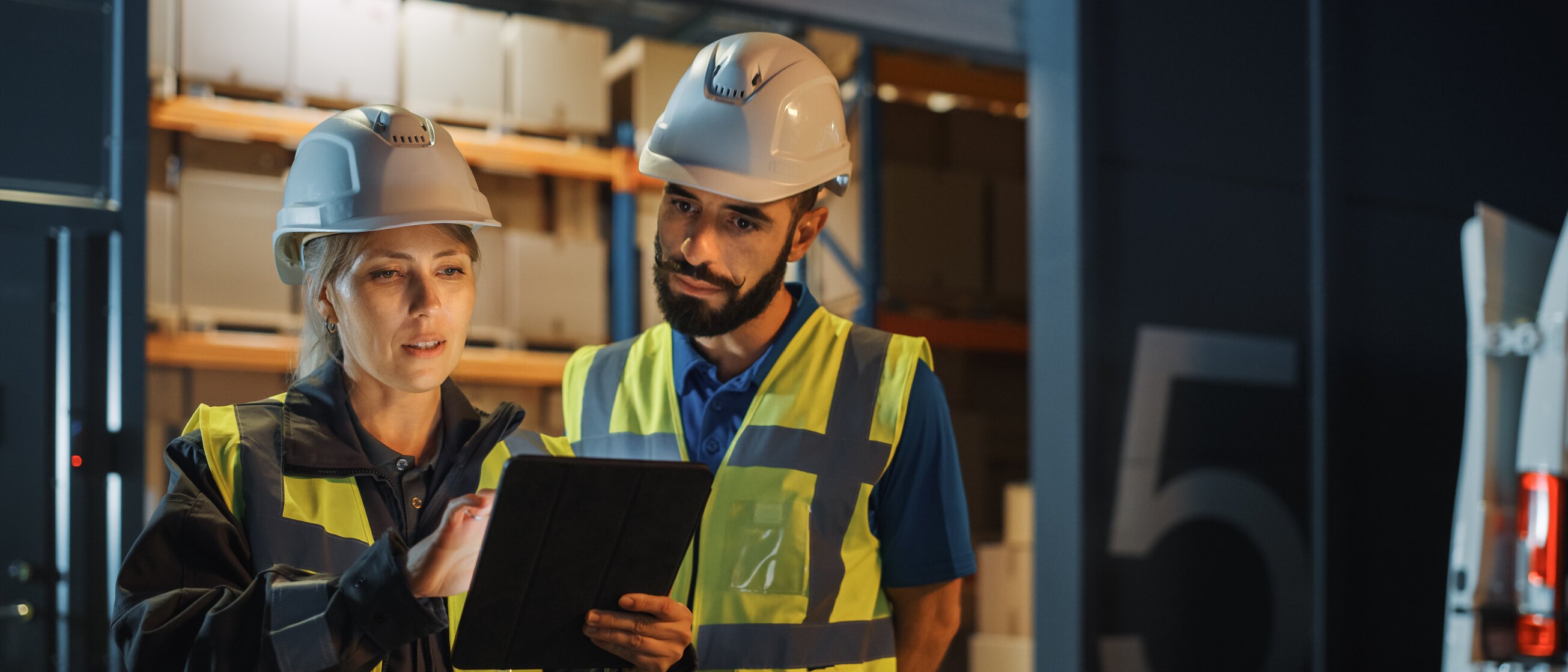 Zwei Logistikfachkräfte mit Schutzhelmen und Warnwesten besprechen anhand eines Tablets Abläufe im Lager – Symbol für moderne Logistik, Digitalisierung und effiziente Prozesssteuerung.
