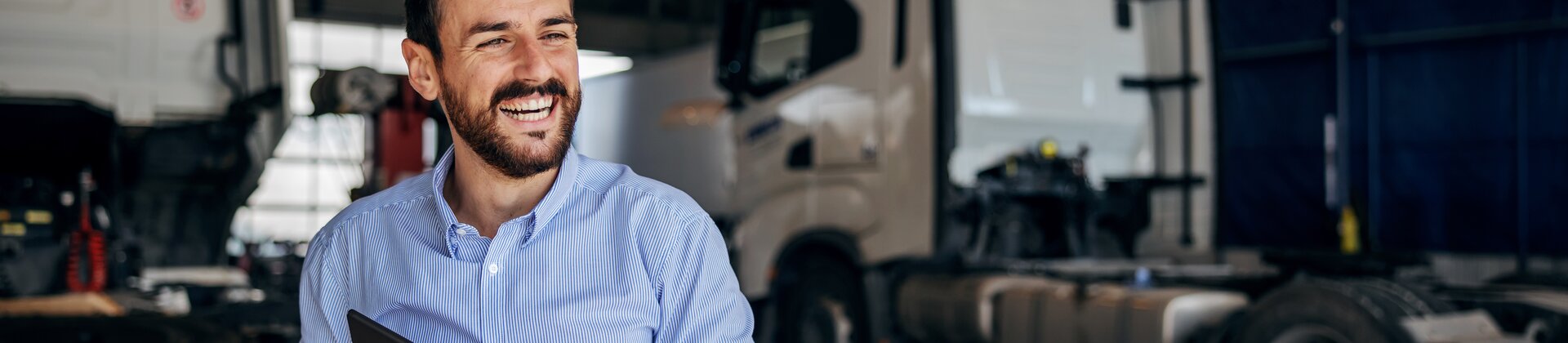 Ein lächelnder Mann steht mit einem Tablet in der Hand in einer Lkw-Werkstatt zwischen mehreren Lastwagen.