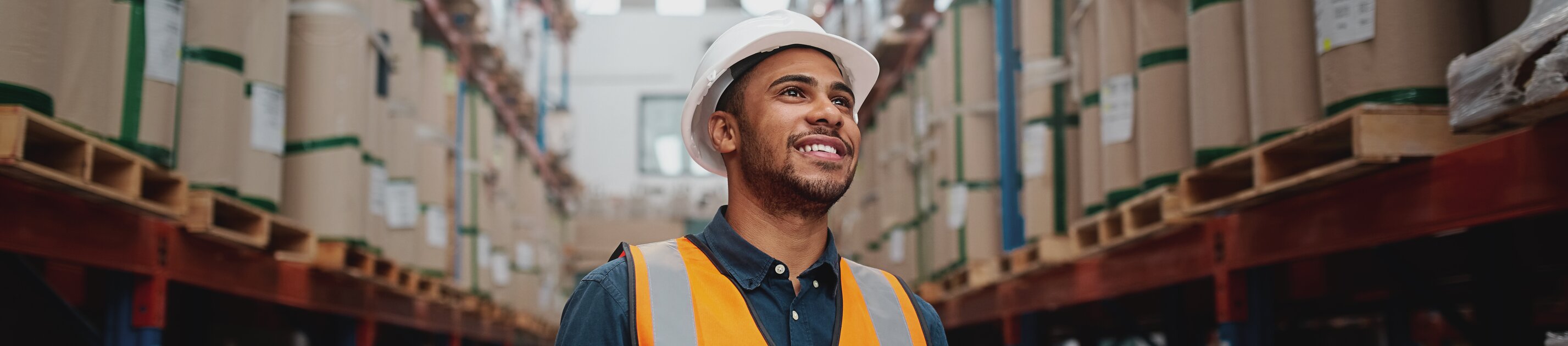 Ein Lagerarbeiter mit Helm und Warnweste prüft Bestände auf einem Tablet zwischen hohen Regalen.