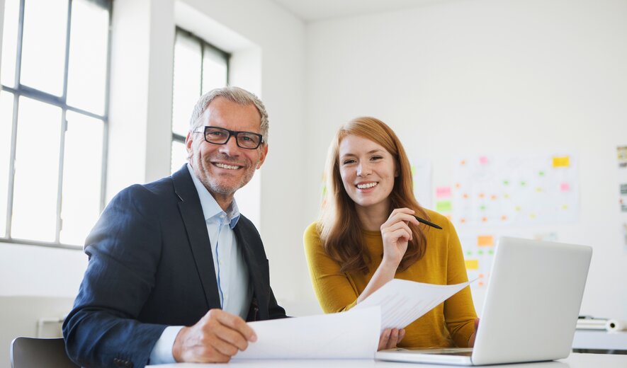 Ein Mann und eine Frau arbeiten gut gelaunt gemeinsam an Unterlagen an einem Schreibtisch.