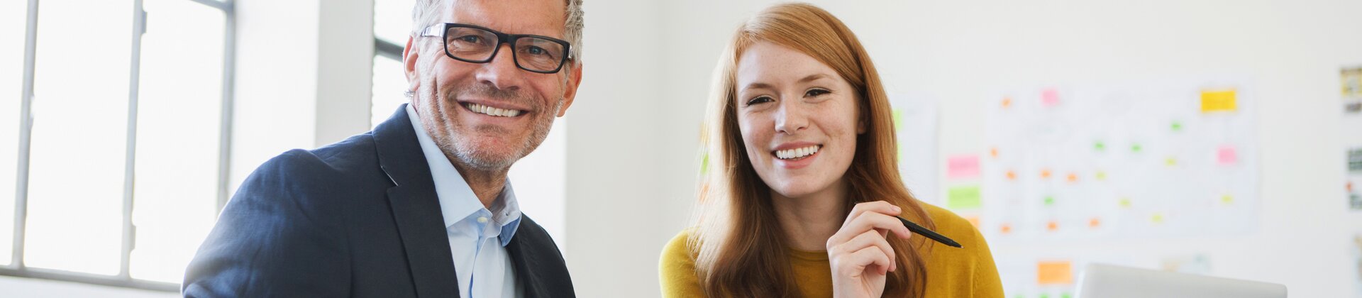 Ein Mann und eine Frau arbeiten gut gelaunt gemeinsam an Unterlagen an einem Schreibtisch.