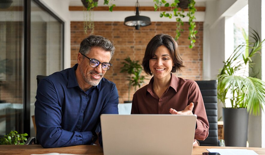 Zwei Personen sitzen lächelnd vor einem Laptop und besprechen gemeinsam etwas am Bildschirm.