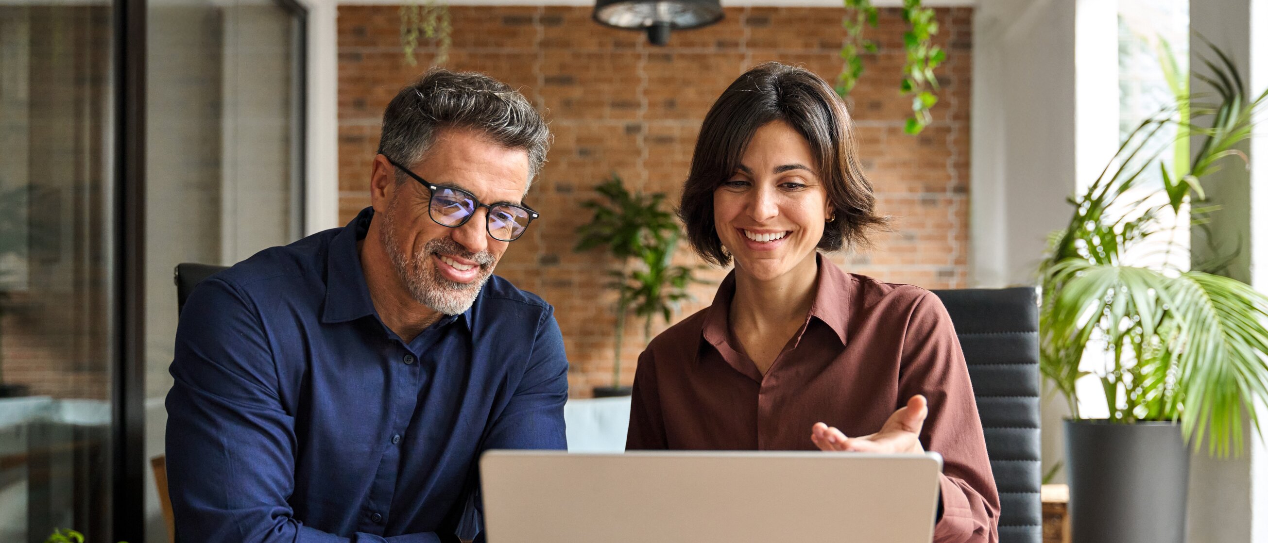 Zwei Personen sitzen lächelnd vor einem Laptop und besprechen gemeinsam etwas am Bildschirm.