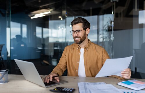 Ein Mann sitzt lächelnd am Schreibtisch mit Laptop und Unterlagen – Symbol für erfolgreiches Lernen, Motivation und praxisorientierte Weiterbildung im kaufmännischen Bereich.