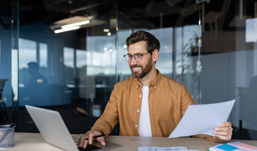 Ein Mann arbeitet lächelnd am Schreibtisch mit Laptop und Unterlagen in einem modernen Büro.