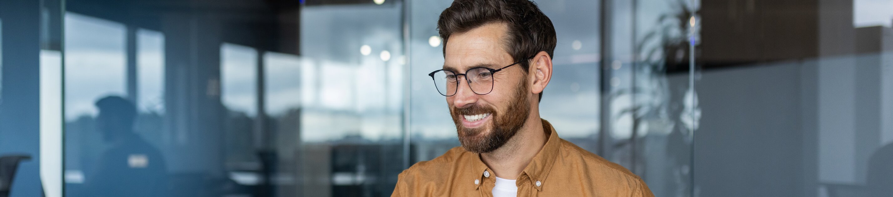 Ein Mann arbeitet lächelnd am Schreibtisch mit Laptop und Unterlagen in einem modernen Büro.