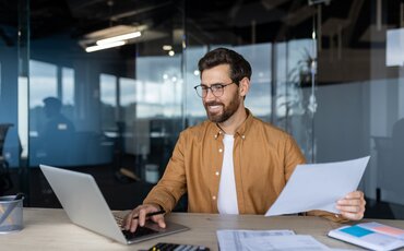 Ein Mann sitzt lächelnd vor Laptop und Unterlagen – Sinnbild für praxisnahes Lernen, technisches Know-how und erfolgreiche Weiterbildung im Management.