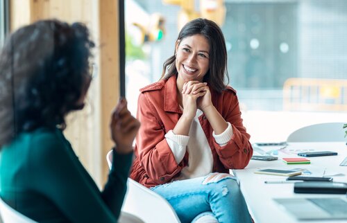 Zwei Frauen sitzen sich in einem Gespräch gegenüber, eine lächelt offen und hört aufmerksam zu – Symbol für erfolgreiche Kommunikation, Empathie und vertrauensvolle Mediation.
