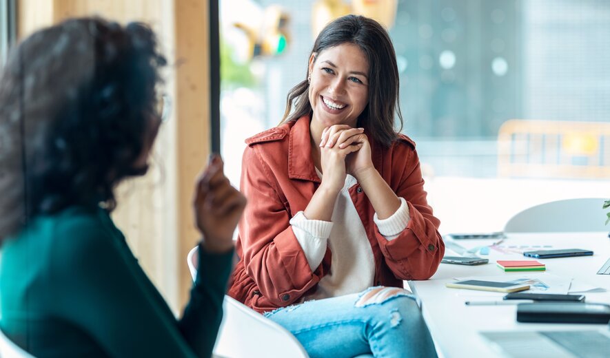 Eine junge Frau sitzt lächelnd in einem Gespräch und wirkt dabei offen und interessiert.