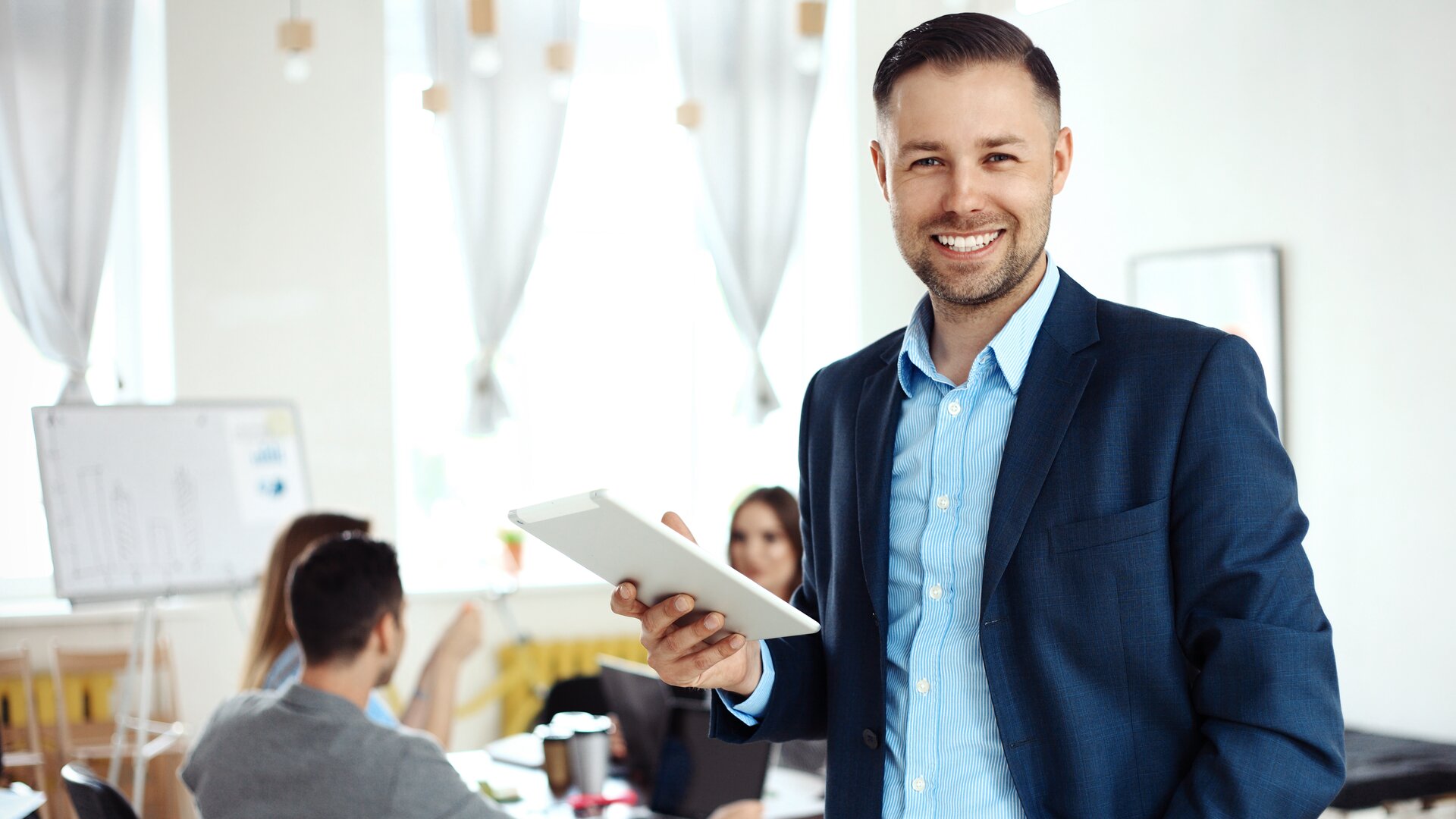 Ein Mann in business-typischer Kleidung steht lächelnd im Vordergrund und hält ein Tablet, während im Hintergrund ein Team an einem Tisch zusammenarbeitet.