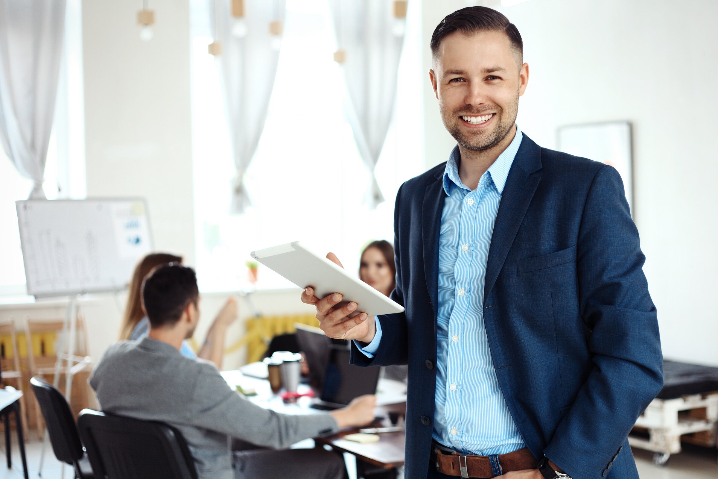 Ein Mann in business-typischer Kleidung steht lächelnd im Vordergrund und hält ein Tablet, während im Hintergrund ein Team an einem Tisch zusammenarbeitet.