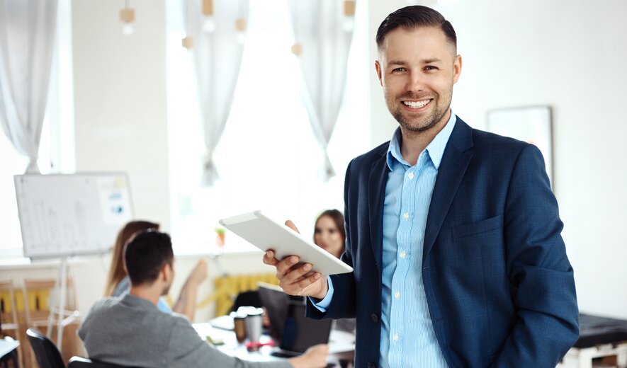 Ein Mann in business-typischer Kleidung steht lächelnd im Vordergrund und hält ein Tablet, während im Hintergrund ein Team an einem Tisch zusammenarbeitet.