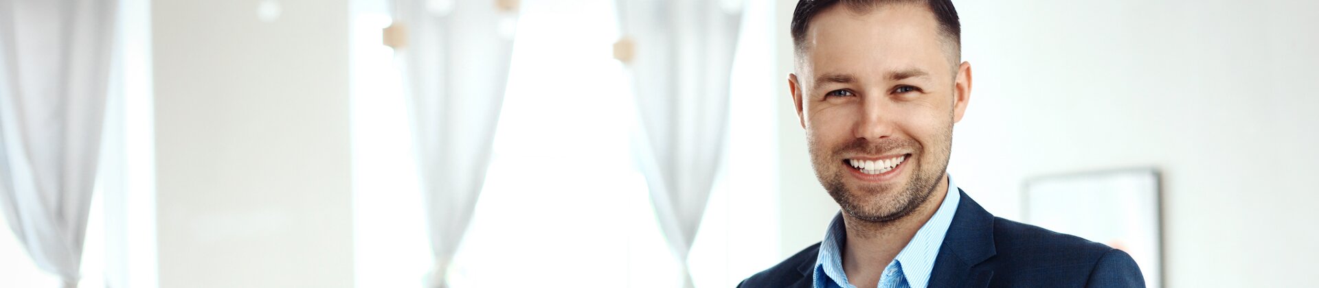 Ein Mann in business-typischer Kleidung steht lächelnd im Vordergrund und hält ein Tablet, während im Hintergrund ein Team an einem Tisch zusammenarbeitet.