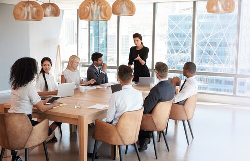 Eine Frau steht am Kopf eines großen Konferenztisches und spricht zu einer Gruppe von Geschäftsleuten, die aufmerksam zuhören.