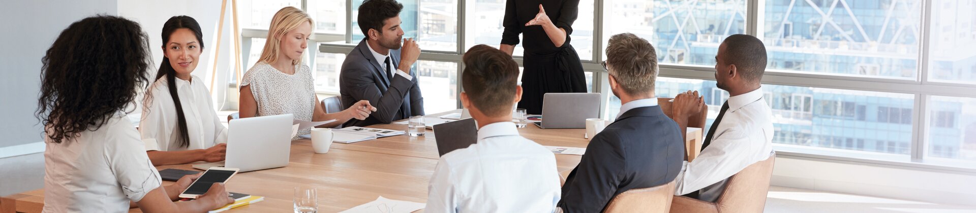 Eine Frau steht am Kopf eines großen Konferenztisches und spricht zu einer Gruppe von Geschäftsleuten, die aufmerksam zuhören.
