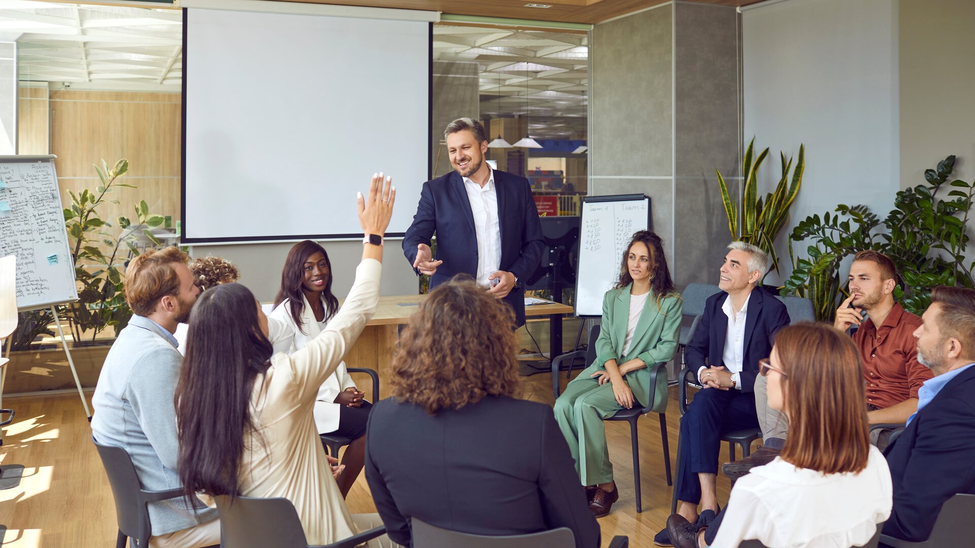 Eine Gruppe von Menschen sitzt in einem Stuhlkreis, während ein Moderator vorne steht und eine Person sich meldet.