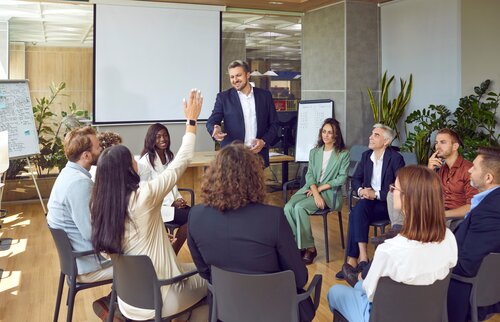 Eine Gruppe von Teilnehmenden sitzt in einem Seminarraum, während ein Moderator vorne spricht und eine Person die Hand hebt – Symbol für aktive Beteiligung, Kommunikation und erfolgreiche Mediation.