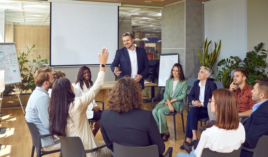 Eine Gruppe von Menschen sitzt in einem Stuhlkreis, während ein Moderator vorne steht und eine Person sich meldet.