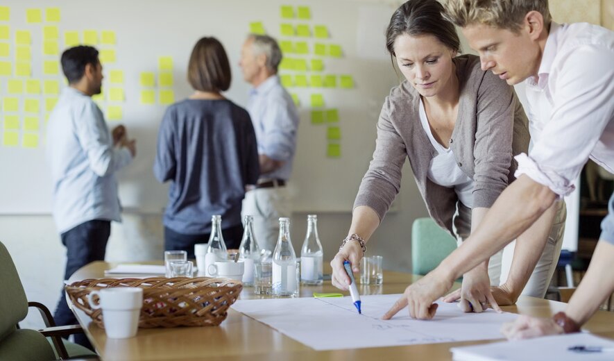 Mehrere Personen arbeiten gemeinsam an Ideen, wobei einige am Tisch skizzieren und andere Post-its an einer Wand anbringen.