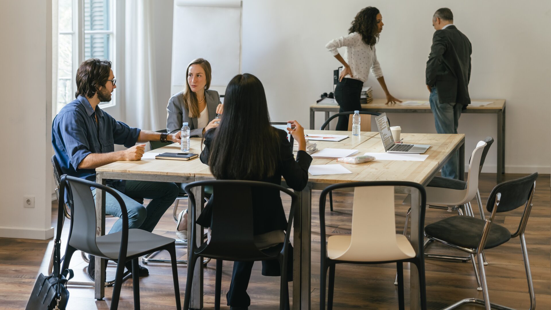 Mehrere Personen sitzen an einem Tisch und besprechen gemeinsam Unterlagen, während zwei weitere im Hintergrund miteinander sprechen.