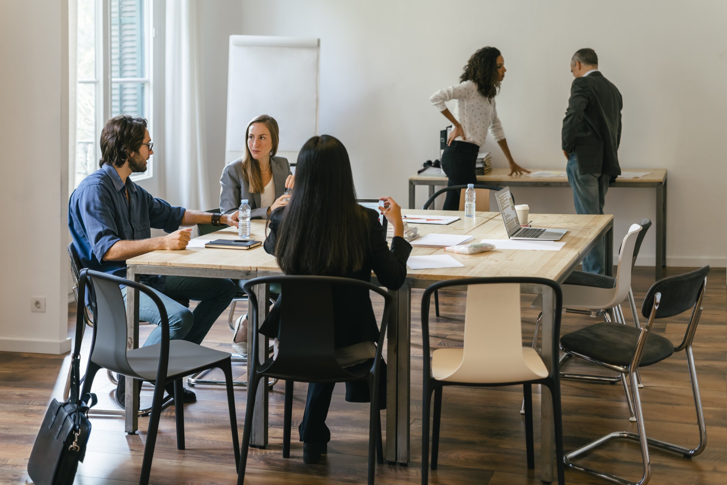 Mehrere Personen sitzen an einem Tisch und besprechen gemeinsam Unterlagen, während zwei weitere im Hintergrund miteinander sprechen.