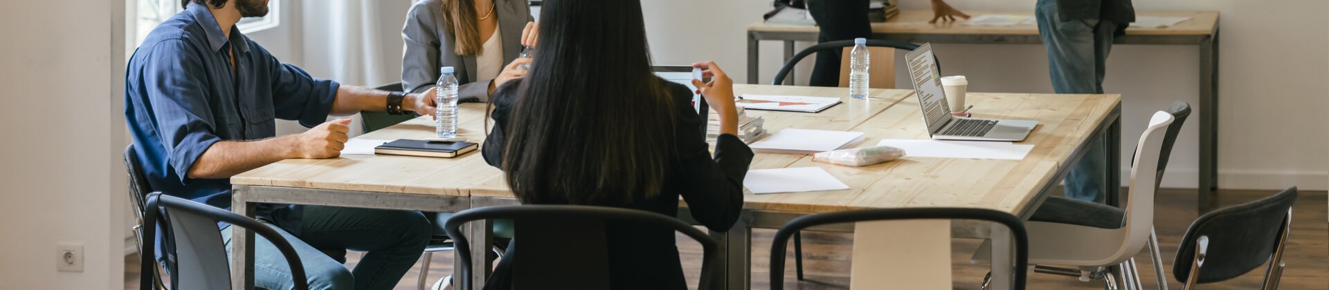 Mehrere Personen sitzen an einem Tisch und besprechen gemeinsam Unterlagen, während zwei weitere im Hintergrund miteinander sprechen.