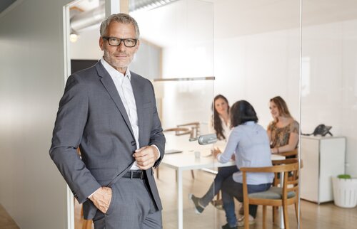 Ein selbstbewusster Mann im Anzug steht vor einer Gruppe im Meetingraum – Sinnbild für erfahrene Trainer, Kompetenzvermittlung und professionelle Ausbildung in Wirtschaftsmediation.