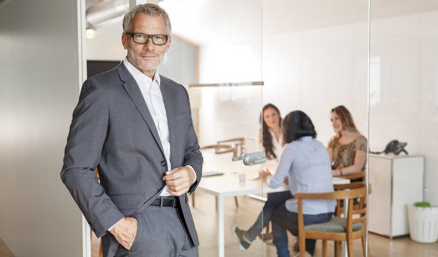 Das Bild zeigt einen selbstbewusst wirkenden Geschäftsmann im Anzug, der vor einem Besprechungsraum steht, in dem mehrere Personen in ein Gespräch vertieft sind.