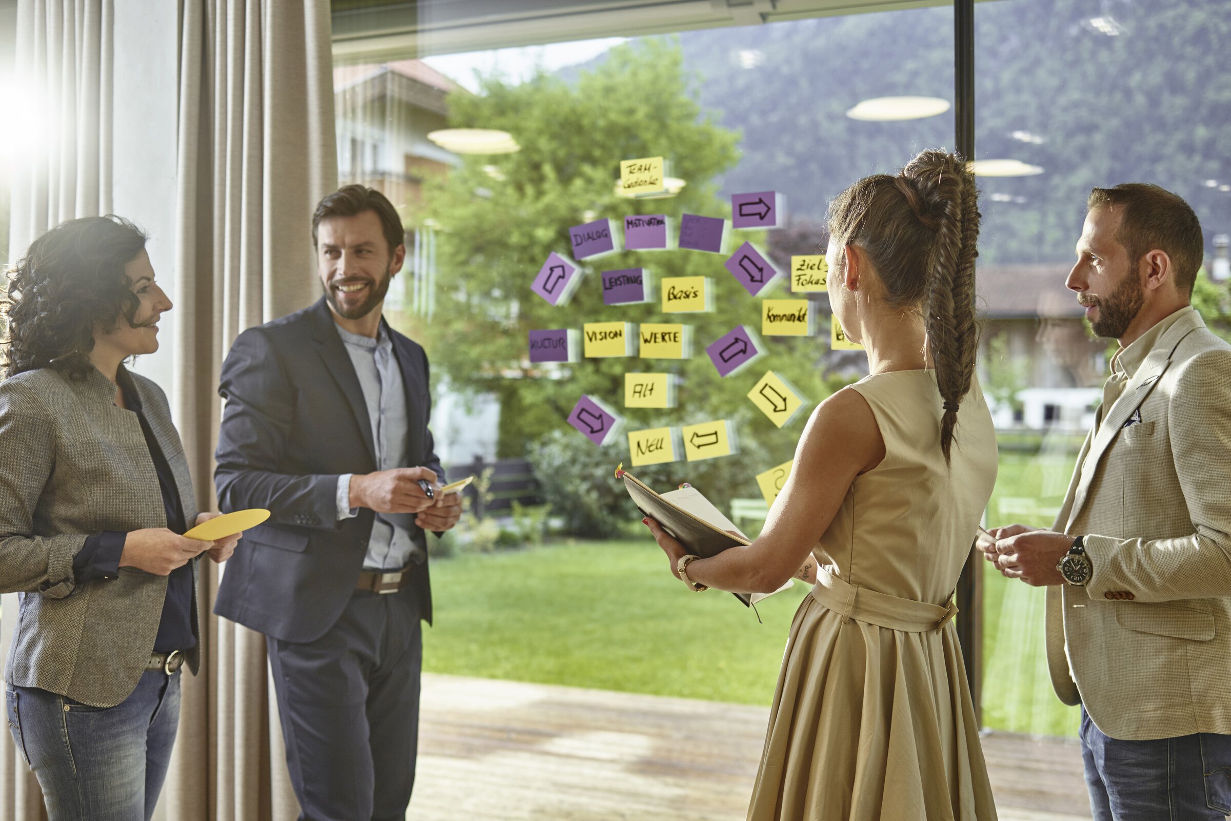 Eine vierköpfige Gruppe hält einen kreativen Workshop vor einer Glaswand voller bunter Haftnotizen.